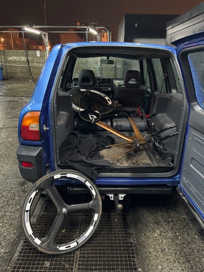 A blue vehicle's rear cargo area is open, revealing a cluttered space. Inside, a bicycle with a light brown frame rests against a grey wall panel, alongside a black bag and a dark cloth. A three-spoke carbon fiber wheel leans against the vehicle's bumper on a wet, metal grated surface. The scene is illuminated by streetlights reflecting off the damp surroundings.