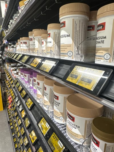 The image shows an aisle in a pharmacy or health food store, likely the Toormina Pharmacy located at 1443 Pacific Highway, Toormina, NSW 2452, Australia. The shelves are stocked with various health supplements, predominantly vitamins and minerals in cylindrical bottles and tubs. Prominent brands visible include Swisse, with several products featuring "Magnesium" and "Magnesium Citrate" on their labels.
The lighting is artificial, typical of an indoor retail environment. The perspective is from eye level, looking down an aisle of shelves. The shelves are organized with products displayed in front of digital price tags, many of which show sale prices. For example, one tag reads "$14.75 Save $14.75" and another displays "$27.50 Save $27.50".
On the lower shelves, there are rows of smaller bottles, some with purple lids, and jars of what appear to be chocolate spread. On the topmost shelf, partially visible, are more bottles with colored lids, including yellow and white. There are no people visible in the image, and the focus is on the product displays and the retail environment. The overall impression is one of a well-stocked health supplement section in a retail store.