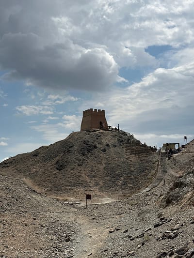 A low-angle shot captures a historic watchtower situated atop a rocky, barren hill under a cloudy sky. The watchtower, constructed of brick, has a crenellated top and an arched entrance. A wooden fence with vertical posts lines a steep staircase that ascends the hill towards the tower. Several people are visible walking up the stairs and near the tower, some are standing near a small, open-sided structure to the right of the stairs. The foreground consists of a rough, rocky path winding through barren terrain, with a small signpost visible in the distance. The scene appears to be a tourist site, likely the Jiayu Pass Great Wall in Jiayuguan, China, given the architectural style of the watchtower and the surrounding arid landscape.