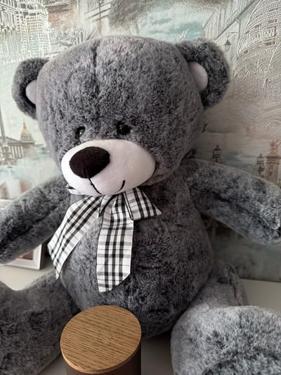 The image shows a close-up of a large, gray, plush teddy bear sitting in front of a wallpaper depicting architectural elements. The teddy bear has black button eyes, a black nose, and a white muzzle with a stitched smile. It is wearing a black and white checkered bow tie. A portion of a wooden cylindrical object is visible in the lower left corner of the frame. The wallpaper behind the teddy bear features a monochromatic scene with buildings and domes, possibly inspired by European architecture. The lighting is bright and even, suggesting an indoor setting. There is no information in the image to suggest the location is in the city of Volzhsky, Russia.