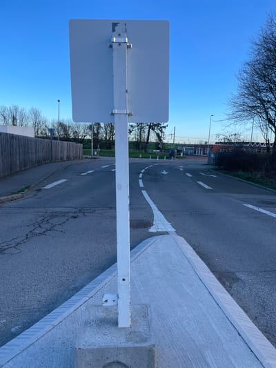 The image displays a street scene in Plaisir, France, viewed from directly behind a plain white, rectangular road sign. This sign is affixed to a white metal pole via two silver-colored clamps. The pole is anchored within a triangular, light grey concrete traffic island that protrudes into the road in the foreground.

The road surface is dark asphalt, exhibiting visible cracking, particularly on the left side of the frame. White painted markings delineate the traffic flow: a solid line bordering the concrete island, dashed lines indicating lanes, and two distinct arrows on the pavement suggesting a left turn and a straight-ahead direction.

To the left, a tall, weathered wooden fence follows the curvature of the road. Beyond the fence, a light-colored building and numerous deciduous trees with bare branches are visible. A streetlight pole stands further down the road on the left. On the right side of the road, a patch of natural ground with similar leafless trees and several white bollards are present.

In the background, a multi-story building with reddish and white sections, potentially a station, is visible. Overhead wires and a bridge-like structure, characteristic of a railway line, are discernible above and behind this building. A solitary, indistinct individual is present in the distant background near the building on the right. The sky above is clear and blue, indicating natural daylight conditions.