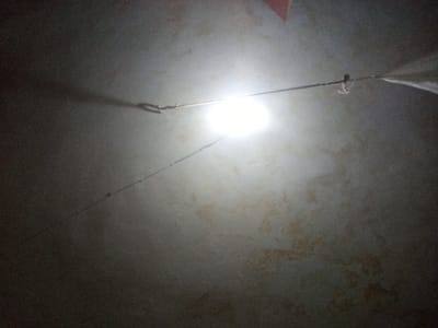 The image depicts a dim room, possibly a bedroom. A bright light source is centrally positioned near the ceiling, casting stark shadows across the textured surface. A thin wire or string stretches horizontally, anchored on either side by hooks. The wire appears to support something, possibly a mosquito net partially visible on the right. The overall ambiance is subdued and somewhat unclear due to the low lighting.