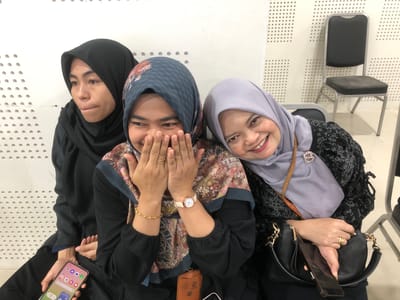 Three women wearing hijabs are sitting side-by-side in what appears to be a waiting area or hallway, with a white wall featuring a perforated pattern behind them. The woman on the left, in a black hijab, looks to the left with her mouth slightly open. The woman in the middle, wearing a patterned hijab, is covering her mouth and nose with her hands, her eyes wide. The woman on the right, in a light purple hijab, is smiling directly at the camera, her head tilted slightly. She is wearing a patterned black top and carrying a black handbag. The woman in the middle is wearing a patterned top with floral motifs and has a watch on her left wrist. The woman on the left is holding a smartphone in her left hand. Two chairs are visible in the background, one black and one gray. The overall setting suggests an indoor public space.