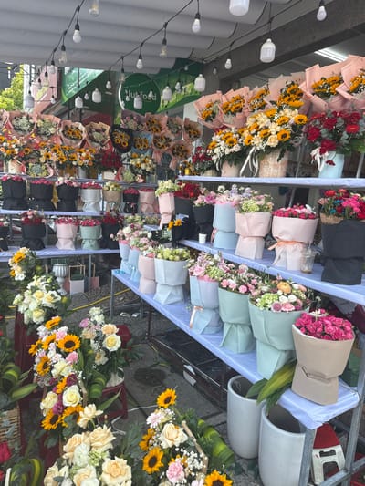 This image captures an outdoor flower stall in Ngũ Hành Sơn Ward, Vietnam, displaying a vast array of pre-arranged bouquets on multi-tiered metal shelving units covered with light-colored fabric. Overhead, a string of bare Edison-style and white spherical light bulbs provides illumination, with a green "clubfood" sign visible in the background. The stall features numerous bouquets, prominently featuring sunflowers mixed with white or yellow roses and filler flowers, often wrapped in peach, pink, light blue, or black paper. Other arrangements include various roses (red, pink, white, yellow), carnations, hydrangeas, and gerbera daisies, wrapped in pastel shades like light pink, light blue, mint green, beige, and black. Some flowers are displayed in white ceramic-like vases or woven baskets on lower levels. The ground is paved, and small red stools along with white cylindrical planters are visible near the base of the display. No people are present in the scene.