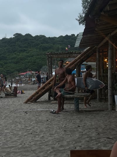 The image shows a sandy beach in Montañita, Ecuador, with coordinates -1.821, -80.756. The scene is dominated by a large wooden structure, likely a restaurant or bar, with a thatched roof and bamboo walls. Three young men are present, all wearing casual clothing. One man sits on a wooden bench with his legs crossed, holding a bottle in his hand. Another man stands next to him, shirtless, and a third man is walking away with a surfboard under his arm. The setting is a beach in the town of Montañita. There are multiple palm trees in the background and a lush green hillside beyond. The sky is overcast with a gray hue. The beach appears to be in a relaxed, post-sunset setting. The people in the scene are interacting casually with each other, appearing to be enjoying a break from the beach. The scene suggests a typical day in Montañita, a popular tourist destination known for its relaxed atmosphere and surfing culture.