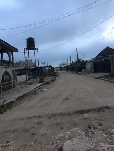 A narrow dirt road stretches into the distance, lined with simple structures and buildings. A water tank towers above a building, adding to the informal landscape. Power lines crisscross overhead. The sky is overcast, creating a somber mood. The environment appears impoverished, with a mix of concrete and natural materials. The overall scene depicts a humble neighborhood or community, typical of a developing area.
