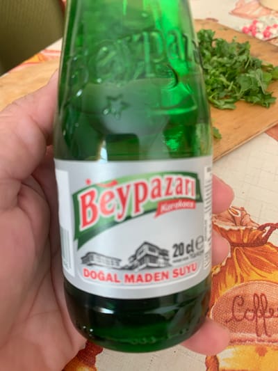 The image shows a person holding a green glass bottle of sparkling water. The bottle has a white label with red and green text and graphics. Visible text on the label includes "Beypazarı", "karadut", "20 cl e", and "DOĞAL MADEN SUYU". The bottle is held horizontally in the person's hands. In the background, a wooden cutting board with chopped green herbs and a decorative tablecloth with a floral pattern are visible. The scene appears to be indoors, possibly in a kitchen or dining area. The lighting suggests it could be daytime. There are no explicit cues about the time of day or weather, but the overall impression is casual and domestic. The image does not convey any specific emotions.
