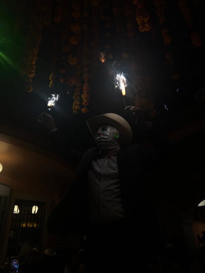 The image depicts an individual in a dark, interior setting within Guanajuato City, Mexico. The subject, viewed from the chest up, wears a light-colored cowboy hat and a metallic, reflective skull-face mask covering the entirety of their face. They are dressed in a dark suit jacket over a lighter-colored collared shirt. Both arms are raised, with each hand holding an active sparkler emitting bright white and yellow sparks. Overhead, numerous garlands composed of orange-yellow flowers, consistent with marigolds, are suspended from the ceiling. A green light source or beam is visible in the upper-left quadrant of the frame. In the lower-left background, architectural elements include a wall-mounted spherical light fixture and a dark opening resembling a barred window or doorway.