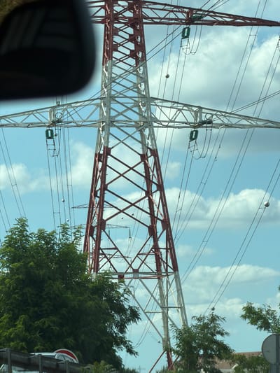 This is a photograph of a large metal power line pylon against a sky with white clouds. The pylon is constructed from red and white latticed metal beams. Multiple power lines are suspended from the pylon, stretching across the frame. Small spherical objects, likely dampers or markers, are visible on some of the power lines. In the lower left corner, a portion of a dark, curved object, possibly a car's rearview mirror, is visible. On the right side of the image, green trees are present, and a red and white traffic sign is partially visible. The sky suggests it is daytime. The orientation of the image is vertical.