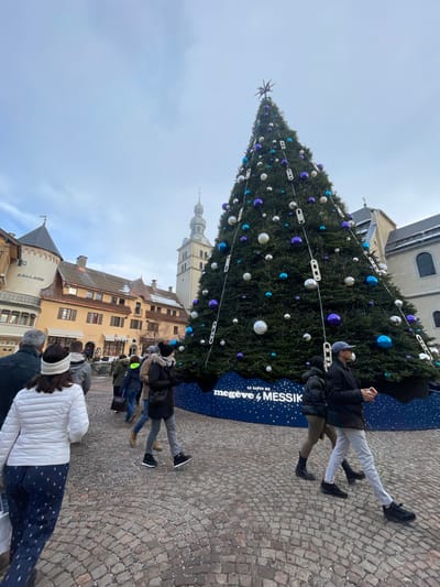 The image displays an outdoor public square in Megève, France, under an overcast sky. A large, conical Christmas tree, decorated with numerous spherical ornaments in silver, blue, and purple, and vertical white decorative elements, occupies the center-right. A star-shaped topper adorns the tree's peak. The tree's base is encircled by a dark blue banner with white snowflake patterns and the text "LE SAPIN DE megève & MESSIK."

To the left of the tree, a row of traditional alpine-style buildings with light-colored facades, multiple stories, and sloped roofs is visible. The leftmost building features a small turret and a sign reading "A. GALLARD." In the center-background, a tall, light-colored bell tower with a distinct spire rises above the other structures. A darker, stone-faced building is partially visible to the immediate right of the bell tower.

The ground is paved with cobblestones laid in a radial pattern, with scattered white debris or light snow visible across its surface. Approximately twelve individuals are present, walking in various directions across the square. They are dressed in winter attire, including heavy coats and hats. In the immediate foreground, a person wearing a white puffer jacket and dark patterned pants walks away from the camera. On the right side, an individual in a dark jacket, light trousers, and a baseball cap walks left-to-right, holding a smartphone. No direct interactions between individuals are discernible.
