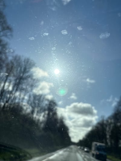 The image captures a forward-facing view from inside a vehicle, looking through a wet and dirty windshield. Numerous water spots, dust, and several distinct, irregular, whitish-clear smudges are visible across the glass, particularly prominent in the upper half. The bright sun is positioned in the upper-middle left, appearing as an intense white light source with a distinct green lens flare below it. The sky is light blue with scattered white cumulus clouds. Leafless trees line both sides of a wet asphalt road that recedes into the distance, creating an avenue effect. Several vehicles are observed ahead on the road, appearing blurred; a white truck or van is discernible on the right side, followed by a darker vehicle. The scene is located in West Felton, United Kingdom.