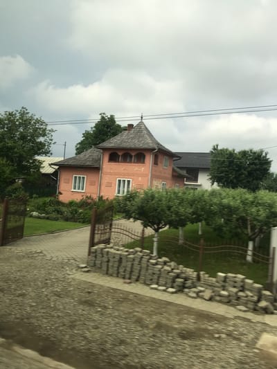 This image depicts a residential scene in Vicovu de Sus, Romania, under an overcast sky. The central focus is a two-story peach-colored house featuring a distinctive dark gray shingled roof with a prominent, almost conical turret-like structure on its left side. The house displays multiple white-framed windows, including three on the ground floor facade and an arched loggia or balcony with four openings on the upper floor. The facade exhibits subtle decorative patterns. To the right and slightly behind, a newer section of the house or an adjacent building with a dark gray roof and white walls is partially visible.

In the foreground, an open set of dark brown ornamental metal gates leads to a paved driveway made of light gray interlocking pavers. The immediate foreground consists of a coarse, dark gray gravel or dirt surface, possibly part of a road or unpaved area. To the right of the driveway entrance, a stack of light gray concrete pavers or bricks is neatly arranged along a low, dark brown metal fence that borders a well-maintained green lawn. A garden with various green shrubs and some discernible red flowering plants is situated between the driveway and the house. A line of small, neatly trimmed trees with white-painted trunks runs along the fence line on the right side of the property. Numerous green trees are visible around and behind the houses, contributing to a lush environment. Overhead, dark utility lines traverse the upper portion of the frame against a sky filled with light and darker gray clouds. No individuals or animals are present in the scene.