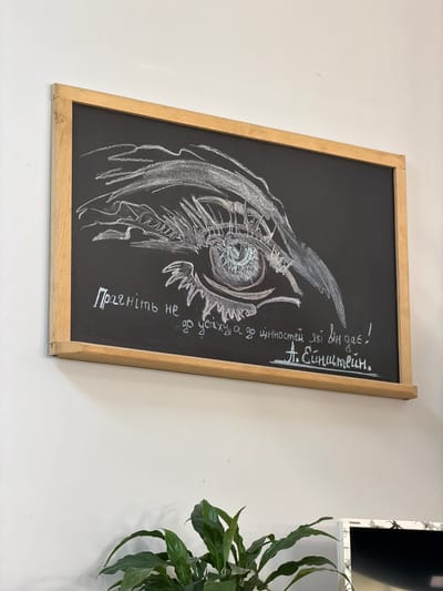 A chalk drawing of an owl's eye on a blackboard is displayed on a white wall. The drawing is rendered in white and light pink chalk, with intricate details in the owl's eye. To the right of the drawing, white chalk writing in Ukrainian is visible. The text reads, "Прагніть не до успіху, а до цінностей які він дає! А. Ейнштейн." which translates to "Strive not for success, but for the values it gives! A. Einstein." The blackboard is framed by a light-colored wooden border. To the right of the blackboard, a corner of a black electronic device with the brand name "TOSHIBA" visible is mounted on the wall. A lush green plant with broad leaves is positioned in the lower right corner of the frame, partially obscuring the wall and the electronic device. The scene appears to be indoors, possibly in a classroom or creative space, given the blackboard and the artistic rendering. The lighting suggests daytime.