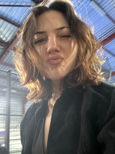 A close-up selfie, rotated 90 degrees clockwise, features a young woman with brown, wavy hair. Her face is illuminated by bright natural light, creating highlights on her hair and skin, and casting subtle shadows. Her eyes are mostly closed or squinting, and her lips are playfully pursed, suggesting a kiss or a "duck face" expression. She is wearing a dark, likely black, jacket or top.
The background indicates an indoor setting, possibly a large industrial building, warehouse, or gymnasium, given the visible high ceiling constructed with blue corrugated panels and metal support beams. The bright lighting suggests it is daytime. The woman's expression conveys a playful and lighthearted mood. No text is visible in the image. The scene is located in Eckbolsheim, France.