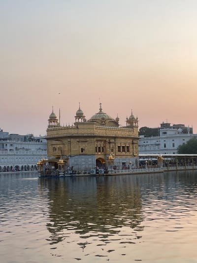 The image shows the Golden Temple in Amritsar, India. It is a Sikh gurdwara, and is considered the holiest site in Sikhism. The temple is made of gold and is surrounded by a large pool of water. The temple is illuminated by the soft light of the setting sun. There are people on the walkway surrounding the temple, and some are in boats on the water. The sky is a gradient of pale orange and pink, with some faint clouds. The water reflects the golden colors of the temple and the sky. The buildings in the background are white with arches.