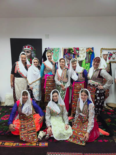 In Ribnovo, Bulgaria, a group of eleven women are pictured wearing traditional Bulgarian folk costumes. The women are arranged in two rows, with the front row kneeling and the back row standing. The costumes are richly adorned with colorful embroidery, beads, and sequins, featuring white headscarves, vests over patterned blouses, and long, brightly colored skirts. The backdrop includes a large framed portrait of a woman in traditional headdress, and display racks with more folk costumes and decorative textiles. The floor is covered with intricately patterned rugs. The scene suggests a cultural event or celebration in Ribnovo.