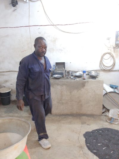An interior scene in Olomoda, Nigeria, depicts a workspace or processing facility. A dark-skinned adult male, wearing a blue long-sleeved shirt and matching trousers with white crocs, walks towards the viewer from the center-left, maintaining eye contact with the camera. His shirt is partially unbuttoned.
The environment features light-colored, possibly concrete, walls with exposed electrical conduits and wires running horizontally. In the upper left, a wall-mounted light fixture or fan is visible. To the left foreground, a large, light-colored metal basin rests on a red and green support. Behind the man, to his left, two black buckets are stacked.
On the right side of the image, a concrete block structure or bench serves as a workstation. Upon it are several silver metallic bowls, a clear plastic water bottle, a blue spray can, and what appears to be a compact scale or analytical device along with various small tools and components. Behind the bench, a coiled off-white hose or cable hangs on the wall, near exposed electrical components.
The concrete floor shows a dark, granular material, possibly seeds or charcoal, spilled in a irregular pile on the right side. Nearby, a large circular sieve or screen with a wooden handle, a plastic bag containing unidentified items, and a blue-handled tool are present. The overall lighting suggests an indoor setting, likely from overhead fixtures.