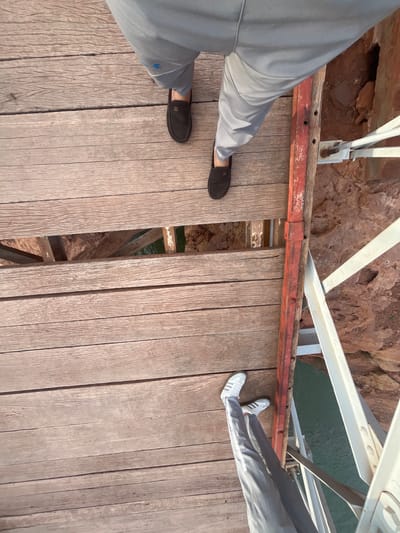 View from above of a wooden bridge in Tabaroucht, Morocco. A person wearing white Adidas sneakers and grey pants stands on the left side of the bridge. A person wearing black slip-on shoes and grey pants stands on the right side. The bridge has gaps between the wooden planks, revealing the rock and water below. The bridge is supported by a white metal structure.
