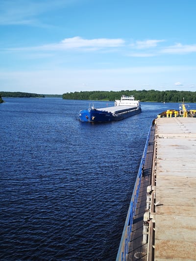 The image depicts a bright, sunny day on a wide river in Nova, Russia. The water is a deep blue and shows small ripples, indicating a gentle breeze. The scene is dominated by two barges. In the foreground, a large, flat-top barge occupies the right side of the frame, its weathered deck stretching towards the viewer. A blue railing runs along its left edge, separating it from the water.
In the mid-ground, a second, larger barge is sailing away from the viewer. This barge is dark blue and appears to be carrying a superstructure or cargo area towards the stern. The name "ГЕCKAP" is clearly visible in white lettering on the side of this barge.
The riverbanks are lined with dense, dark green forest, suggesting a natural, undeveloped landscape. The sky is a vibrant blue, with scattered, wispy white clouds adding depth to the scene. The lighting indicates it is likely midday, given the bright sun and clear visibility. The overall atmosphere is one of peaceful navigation on a calm summer day. There are no people visible in the image.