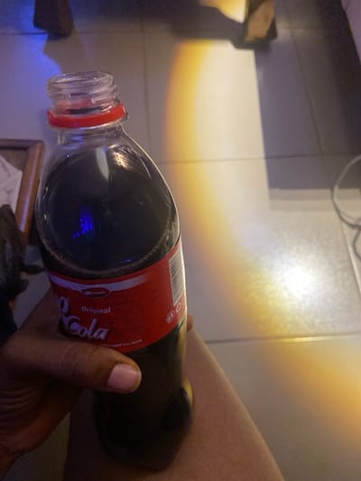 A person holds a plastic bottle of cola in their hand. The bottle is partially filled with a dark liquid. The label is red with white lettering. The background shows a tiled surface with a light reflection creating a gradient. An out-of-focus object is visible below the bottle. The scene is indoors with artificial lighting.