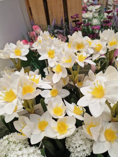 This is a close-up, slightly angled photograph of a collection of artificial flowers. The primary subjects are white flowers with bright yellow centers, resembling daffodils or narcissus. They are arranged in a dense cluster, with many individual blooms in various stages of openness. Interspersed among them are smaller clusters of tiny white flowers. In the background, out of focus, are other artificial flowers in shades of pink and purple, and a section of a wooden crate. Some of the flowers have small paper tags attached to their stems, with visible text including "IMPORTAN", "MATION", and "ern&Fable". The lighting appears to be artificial, suggesting an indoor setting, possibly a retail store or a display. There are no people or animals visible. The overall impression is a bright and colorful display of decorative floral elements.
