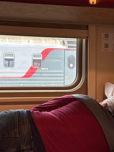 The image shows a person asleep on a train. The person is lying on their side with their back to the camera. They are wearing a red shirt and blue jeans. Outside the window, another train is visible. The scene takes place on a train traveling through Kotelnich, Russia.