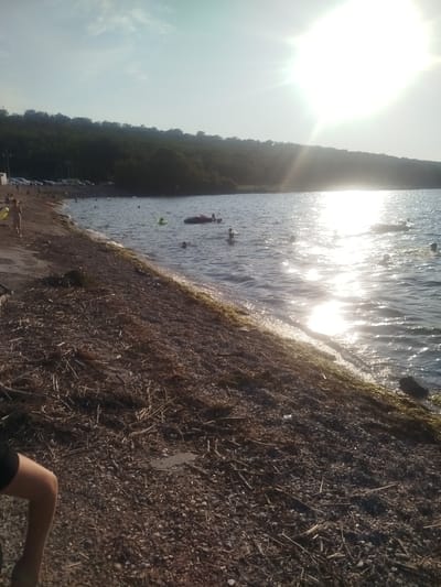 This image depicts a lively beach scene on a sunny day, likely during the late afternoon given the low angle of the sun and its strong glare on the water. The setting is a shoreline with a pebble and driftwood-covered beach bordering a body of water. In the background, a dense, tree-covered hillside rises, with a parking lot visible at its base, suggesting a public access area. The water is dotted with several people swimming, some with inflatable rings, and a larger inflatable raft is also present. The sun's intense reflection creates a bright, almost hazy effect across the water's surface, indicating fair weather. The overall mood appears to be relaxed and recreational, with individuals enjoying the water and the warm sunlight. The presence of cars in the background and numerous people in the water points to a popular, accessible beach location. No visible text is present in the image.