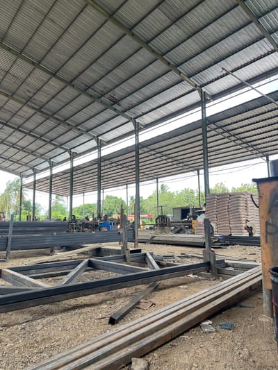 An open-sided industrial shed with a corrugated metal roof supported by steel beams and columns, located in the city of Lambarobiluy, Indonesia. The ground is covered with gravel and dirt, and the shed is filled with construction materials, primarily long metal bars stacked in piles and pre-fabricated steel frames. In the background, there are stacks of cement bags, industrial machinery, and some vehicles, including a red forklift. Several palm trees and other foliage are visible outside the shed, indicating a tropical or subtropical climate. There are a few people present, some of whom appear to be workers. The overall scene depicts an active construction or fabrication yard.