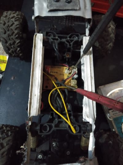 An overhead forensic view captures the underside of a remote-controlled off-road vehicle, likely in a state of repair or inspection. The vehicle features a dark plastic chassis, with two large, heavily-treaded black tires visible at the outer edges. Two coil springs, part of the suspension system, are also discernible. Running longitudinally along the chassis are two light-colored, possibly wooden or plastic, support strips. Internally, a central yellow circuit board or electronic module is exposed, featuring various connected wires, including prominent black, red, and yellow conductors. A black-handled screwdriver with a metallic shaft is positioned with its tip near a small white connector on the yellow module, indicating manipulation or adjustment. To the right, a red test lead, likely from a multimeter, points towards the internal electronics, with a partial view of a white-cased device, possibly a "HELP MODEL SM" multimeter, visible in the lower right background. The immediate environment is dark and indistinct, suggesting an indoor workspace. No specific visual information within the image confirms a precise location within Mornington, Australia.