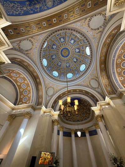This is a low-angle, wide shot of the ornate interior of a domed building, likely a public building or place of worship. The architecture features intricate golden decorative elements on a cream-colored background, interspersed with panels of blue and gold geometric patterns and floral motifs.
Several large, white columns with golden Corinthian capitals extend upwards, supporting the domed ceiling. The ceiling itself is a complex structure with multiple smaller domes and arched sections, all adorned with detailed artwork. In the upper part of the image, a large chandelier with multiple yellow lights hangs from the ceiling.
Towards the right side of the image, a flat-screen television is mounted on the wall, displaying a colorful image, possibly of a stained-glass window or artwork. The lighting appears to be artificial, with light also entering from some small, oval windows in the ceiling. The overall impression is one of grandeur and opulence.
The location context provided is "посёлок дачного хозяйства «Архангельское», Russia," suggesting the building is located in this area. There is no visible activity or event taking place, and no discernible text in the image. The time of day is not indicated by natural light.