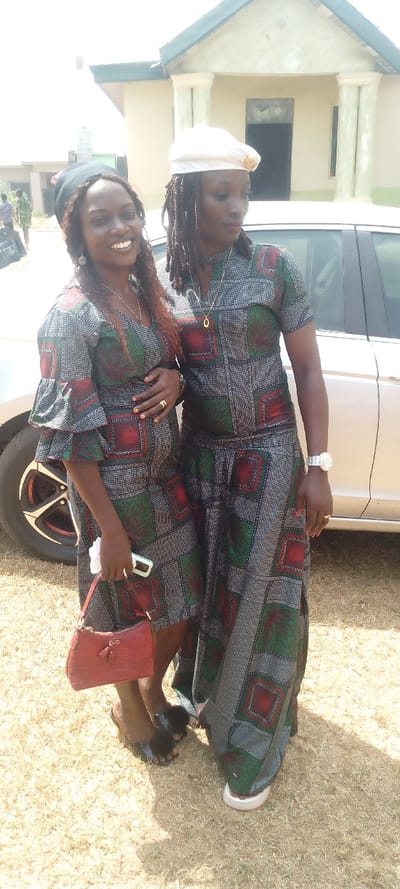Two women stand side-by-side in matching patterned dresses made from green, red, and gray fabric. The woman on the left wears a black head covering and holds a red purse. The woman on the right wears a white beret. Both wear necklaces. They are standing on dry grass with a car and a building in the background. People are walking in the distance.