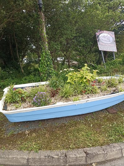 The image shows a blue and white boat repurposed as a planter, filled with various plants and flowers. The boat is situated on a gravelly surface next to a grassy area with a concrete curb in the foreground. Behind the boat, a wooden pole is wrapped in ivy, and a sign for "BONNERS BAR" is visible in the background, indicating a location in Mullaghshouff, likely near Annagry West, Ireland, as suggested by the text on the sign. The environment appears to be outdoors, with trees and foliage surrounding the scene. The lighting suggests it is daytime, with an overcast sky. No people are present in the image.