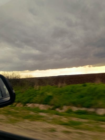 The image depicts a rural landscape near Byal Izvor, Bulgaria, viewed from inside a moving vehicle. In the foreground, the upper left quadrant of a black car rearview mirror is visible, partially obscuring the lower left corner. Below the mirror, a dark horizontal strip represents the car's window frame. The immediate mid-ground exhibits significant motion blur, showing patches of light brown ground interspersed with strips of vibrant green vegetation, indicating movement relative to the scene. Further into the mid-ground, a wide horizontal band of dark brown, freshly plowed earth dominates, suggesting an agricultural field. This field extends towards a low, dark horizon. Above the horizon, a distinct band of bright golden-yellow light, likely from the sun, is visible. Above this light band, the sky is predominantly covered with dense, textured grey clouds, filling the upper two-thirds of the frame. Faint vertical structures, potentially power line towers, are discernible on the distant horizon against the bright light band. No individuals are present in the scene.