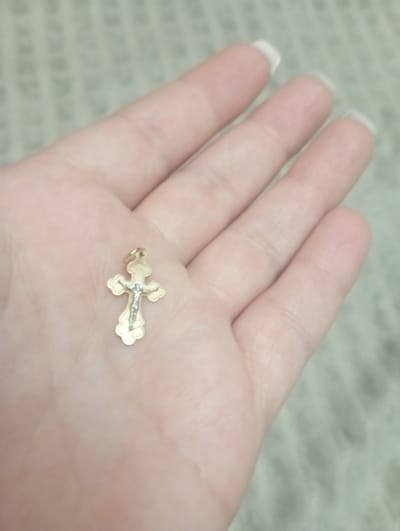 This media file shows a close-up, top-down view of a hand holding a small, ornate, gold-colored Christian cross pendant. The hand is open, with the fingers slightly curled, and the palm is visible. The cross is positioned near the center of the palm, resting on the skin. The pendant appears to be detailed, with intricate carvings and possibly some embedded stones or enamel work visible on its surface, particularly on the center of the cross. The hand has well-maintained fingernails with a French manicure. The background is a textured, light-colored surface, possibly fabric or paper, with a subtle pattern that appears to be text in a foreign language, likely Cyrillic script, which is out of focus. The lighting is soft, suggesting an indoor setting or overcast daylight. The overall impression is one of personal reverence or contemplation. There are no explicit indications of time of day, weather, or strong emotions, but the gentle presentation of the pendant in the hand suggests a moment of quiet reflection or display. The image does not contain any discernible text that can be read clearly.