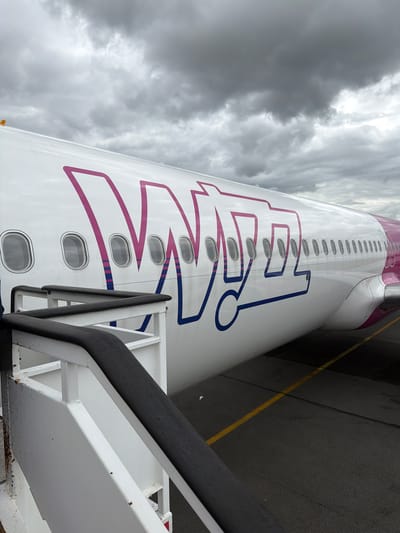 The image captures a close-up view of the side of a Wizz Air airplane, parked on an airport tarmac, likely at Beauvais-Tillé Airport in Tillé, France. The aircraft's white fuselage is prominently displayed, featuring the distinctive "WIZZ" logo in stylized pink and blue lettering that spans a significant portion of its side. A continuous line of oval passenger windows runs along the fuselage, with several visible beneath the text.

In the foreground, an aircraft boarding stair is positioned against the plane, with its black handrail and white structural frame visible, suggesting active boarding or deplaning operations. The tarmac below the aircraft features yellow ground markings. The sky is heavily overcast with dense, grey clouds, indicating cloudy weather during daytime. No people are visible in the scene. The perspective is from ground level, looking upwards and along the length of the aircraft's side.