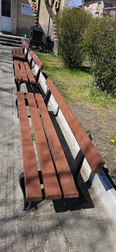 The image shows a row of brown wooden benches on a paved surface in Slatino, Bulgaria. A bush and some grass are visible behind the benches. A building with a sign and steps is to the left. The day is sunny and shadows from the benches can be seen on the pavement. No people are visible in the image.