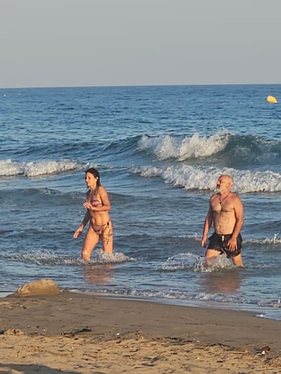 The image depicts two individuals, a woman and a man, wading in the shallow waters of a beach. The woman, on the left, is wearing a patterned bikini and appears to be moving through the water. The man, on the right, is wearing dark swim trunks and has a beard and a necklace. He is also standing in the water. Waves are breaking around them. In the background, the blue sea stretches to the horizon under a pale sky. A yellow buoy is visible in the distance on the right. The foreground shows a sandy beach with some debris. The scene is consistent with a beach in Castelldefels, Spain, known for its long sandy coastline and Mediterranean waters.