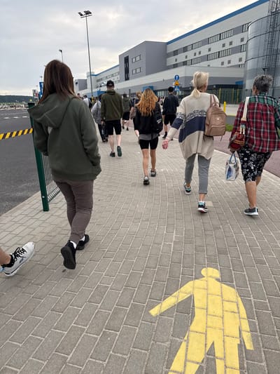 In Chociule, Poland, a group of people walks along a paved sidewalk towards a large modern industrial building. The sky is overcast. The building has a gray facade with blue accents and numerous windows. A yellow pedestrian crossing symbol is painted on the sidewalk. The people are walking away from the camera, and their clothing varies in color and style, including a woman in a green hooded jacket, a man in a green hoodie and shorts, a woman in a patterned cardigan, and another woman in a red and black plaid shirt. Several individuals are carrying bags. The sidewalk is made of grey rectangular pavers.