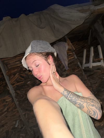 A young Caucasian female is pictured indoors in what appears to be a rustic structure, possibly a banya or sauna, in Maksimovo, Russia. She is wearing a grey and white wool hat and is wrapped in a light green towel. Her eyes are closed, and her head is tilted down, with her right hand touching her cheek. She has visible tattoos on her left arm and shoulder. The background shows wooden planks stacked against a wall, with a draped fabric overhead. The lighting suggests it is nighttime.