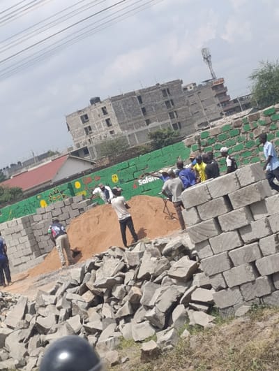 The image displays an outdoor construction scene in Matungulu west ward, Kenya, under an overcast sky traversed by multiple utility lines. In the foreground, a large pile of irregular grey stones and rocks dominates the lower half, interspersed with dry earth and sparse vegetation. A dark, reflective object, possibly a helmet, is partially visible at the bottom center.
The midground features a substantial mound of reddish-brown sand or soil. To the left of the sand, a stack of grey concrete blocks is present. A vertical stack of similar concrete blocks is prominent on the right side of the frame. Several men are actively present: one man in a white t-shirt and dark pants stands on the sand pile, while another, partially obscured, appears to be working with the sand using an implement. A man in a dark patterned shirt and light pants stands near the left stack of blocks. To the right, a group of approximately 5-7 men, some in blue shirts and one in yellow, are gathered near a green wall. Another man in a light blue shirt walks towards the right edge of the frame.
The background includes a green wall adorned with yellow markings and the visible word "Safaricom." Behind this wall, several multi-story buildings, in various stages of construction or appearing unfinished, are visible, characterized by exposed grey concrete and multiple window openings. Some buildings feature black water tanks on their roofs. A prominent cellular communication tower stands against the sky in the upper right background. A red-roofed building is also visible in the far left background.