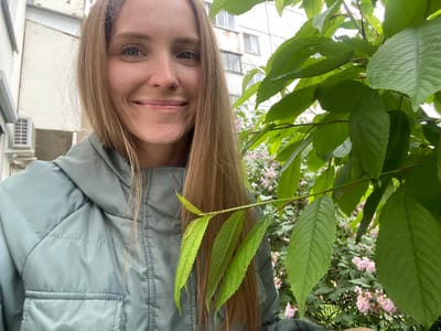 A young woman with long blonde hair is captured in a close-up, outdoor, selfie-style photograph. She is smiling directly at the camera, her face framed by her hair and some green leaves on the right side of the image. She is wearing a light green, puffy jacket with visible stitching and a pocket. In the background, a building with windows and an air conditioning unit is partially visible, along with a bush with small pink flowers. The setting appears to be an urban environment.