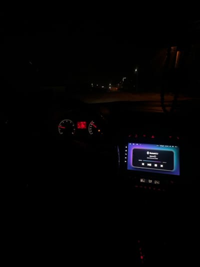 In a dark setting, the interior of a car is visible. A dashboard with a digital display screen shows music playback controls and Bluetooth connectivity. Below it, the car's instrument cluster displays illuminated gauges and a digital odometer. Outside the car window, faint lights of a town or city can be seen, indicating a night drive.