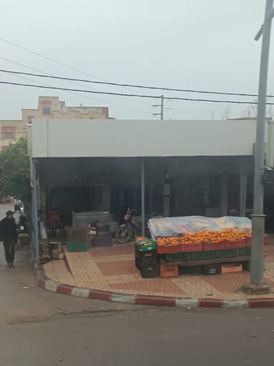 The image depicts a street scene in Oued Jdida, Morocco, under an overcast sky. In the foreground, a dark asphalt road transitions to a patterned sidewalk featuring beige and light brown tiles bordered by a red and white painted curb. On the right side of the sidewalk, a grey utility pole stands. Adjacent to the pole, a makeshift fruit stand displays numerous oranges in stacked red and green plastic crates, with some fruit covered by a transparent plastic sheet. Additional empty crates are visible beneath the display.
Behind the fruit stand, a single-story commercial structure with a flat, light-colored roof extends across the midground. Its facade is recessed, forming an open-front area with a dark interior. Visible inside are several empty green and purple plastic crates, red chairs, and a silver-colored scooter or moped parked to the left of the fruit stand. Further to the left, a dark-colored motorcycle or scooter is partially visible on the sidewalk near the building.
To the far left of the frame, a person wearing dark clothing, including what appears to be a hat, walks from left to right on the sidewalk. A tree with sparse, orange-colored fruit (possibly oranges or tangerines) is situated between this individual and the commercial building. Faintly, another individual is visible further down the left sidewalk.
In the background, multiple-story buildings with light-colored (beige/tan) facades and varying rooflines are present. One notable building features a stepped roof structure with a small, light-blue framed window at its apex. Overhead, multiple dark utility wires span horizontally across the frame, supported by poles visible in the distance. The sky is uniformly light grey, indicating a cloudy day.