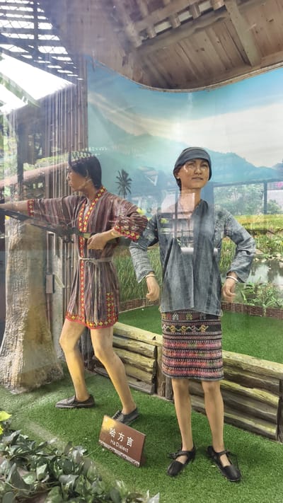This image captures an indoor cultural exhibit in Jiyang District, China, viewed through a reflective glass panel. Two life-sized mannequins, depicting individuals in traditional attire, are positioned on artificial green turf.
The male mannequin on the left stands facing partially right, holding an elongated, dark weapon resembling a crossbow or bow, with his left arm extended forward and right hand near his waist. He wears a knee-length, striped tunic in shades of brown, red, and yellow, adorned with geometric patterns along the borders and cinched with a light-colored sash. He has short, dark hair and dark shoes.
The female mannequin on the right faces forward with a slight turn to the left, displaying a subtle smile. She wears a long-sleeved, grey striped shirt with a V-neck and a knee-length black skirt intricately patterned with colorful geometric designs. She has dark hair covered by a dark cap or headwrap and wears dark, strapped shoes. Each of her wrists bears two silver-colored bangles.
Between the mannequins, a small wooden sign on the turf displays "哈方言" (Ha Fang Yan) and "Ha Dialect" in white text. Behind the figures, a low, simulated log fence extends horizontally, and a large, light-colored vertical element, resembling a tree trunk or pillar, stands behind the male mannequin. The background is a large, curved photographic or painted mural depicting an outdoor landscape with a clear blue sky, distant green mountains, a palm tree, a green field, and a small body of water. Above the backdrop, wooden structural elements of the exhibit ceiling are visible.