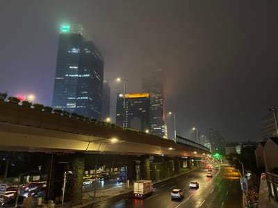 The image depicts a nighttime scene in Jing'an District, China, likely on a street or elevated highway. The atmosphere is hazy or foggy, with limited visibility of distant buildings. A multi-lane road is visible, with several cars and a truck driving on it. The road surface appears wet, reflecting the streetlights and headlights.
In the foreground, a portion of an elevated structure, possibly an overpass or a building walkway, is prominent. This structure is lined with greenery along its top edge and has large, ivy-covered pillars supporting it. Streetlights illuminate the roadway below.
Dominating the background are several tall, modern buildings, characteristic of a city skyline. One of the buildings has a prominent green illuminated logo or sign at its top. Other buildings show illuminated windows. The overall impression is one of urban infrastructure and activity during a foggy night.