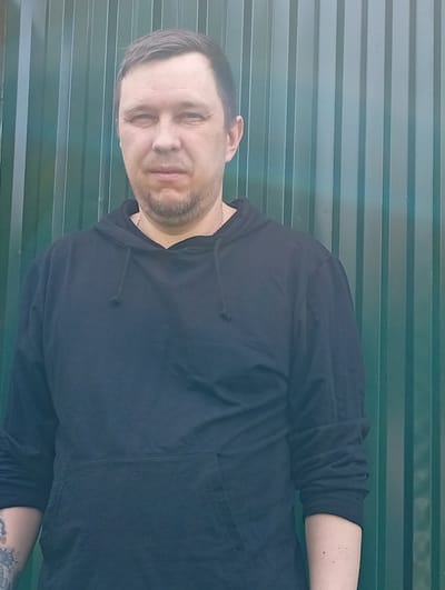 A medium-shot, head-and-shoulders portrait shows a man standing in front of a green corrugated metal fence. The man has short, dark hair, light skin, and a small beard. He is wearing a black hooded sweatshirt with the hood down. A thin chain necklace is visible around his neck. His left arm is visible and shows a tattoo on his forearm. The fence behind him has vertical ridges and is a dark green color. The lighting appears to be natural and overcast. The image was taken outdoors. The location is the city of Pavlovsky Posad, Russia.