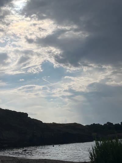A wide shot of a coastal scene in Ahtopol, Bulgaria, captures a dramatic sky filled with a mix of dark, heavy clouds and patches of bright, illuminated sky where the sun is attempting to break through. The sun's rays create a luminous effect through the clouds, casting a diffused light.
In the foreground, a dark, rocky cliff face dominates the left side of the image, its textured surface rendered in silhouette against the brighter sky. The water in front of the cliff is choppy, reflecting the light and giving it a shimmering appearance. There are a few people visible in the water, appearing small against the vastness of the sea and the imposing cliff. To the far right, tall green reeds or grass grow, adding a natural element to the foreground.
Beyond the cliff, a line of dark green trees forms a distant treeline under the cloudy sky. The overall impression is one of a dynamic and slightly moody coastal landscape, typical of the Bulgarian Black Sea coast. The photograph is taken from a low vantage point, possibly from the shore or a beach, emphasizing the scale of the natural elements.