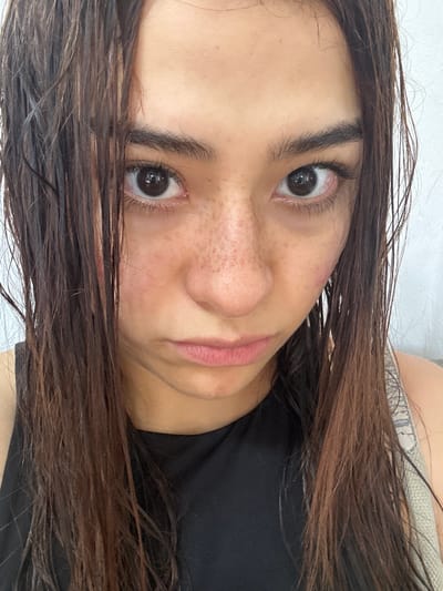 A close-up selfie shows a young woman with dark, wet hair and a pout. She is wearing a black tank top. Her face is directed towards the camera, with her head tilted slightly. Her expression is somber, with her lips pursed and her eyebrows furrowed. She has prominent freckles across her nose and cheeks, and her skin appears flushed, possibly from being in the sun or from the moisture in her hair. The background is a plain white wall, offering no contextual clues about the location. The lighting is bright and appears to be natural, casting soft shadows on her face. No specific landmarks in San Pedro Cholula, Mexico, are visible in the image.