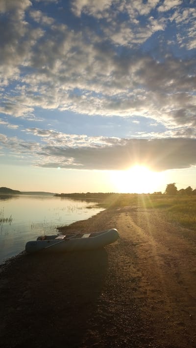 The image displays a serene outdoor scene at sunset or sunrise in Шолья, Russia. The upper portion of the frame is dominated by a clear blue sky interspersed with scattered, illuminated clouds ranging from white to light grey. A brilliant, low-lying sun is prominently positioned on the right side of the horizon, casting intense golden light and distinct rays across the landscape.
Below the sky, a wide, calm body of water, likely a river or lake, occupies the mid-left, reflecting the warm hues of the sky and sun. Sparse aquatic vegetation is visible near the waterline. The opposite bank, a dark, low-lying landmass, is discernible in the distant background.
In the foreground, an inflatable, light grey boat is partially beached on a sandy and gravelly shore that transitions into a dirt path or road. The boat, located in the lower-left quadrant, has what appear to be oars or similar equipment inside. The dirt path runs parallel to the water along the right side of the image, showing subtle tracks and being strongly illuminated by the low sun. To the right of the path is a grassy area leading to distant trees on the horizon. No individuals are visible within the scene.