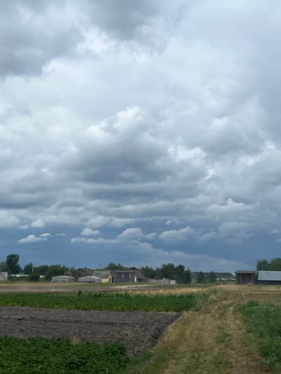 The image depicts a rural landscape in Sobishchytsi, Ukraine, under a dramatic, cloudy sky. In the foreground, there are fields, one with freshly tilled dark earth and another with green crops. A dirt path runs through a field of dry, yellowed grass. In the mid-ground, a small collection of buildings is visible, including houses and agricultural structures like sheds or barns. Trees line the horizon behind the buildings. The sky is filled with heavy, dark gray clouds, suggesting an overcast or possibly stormy day. There are no people or animals visible in the scene.